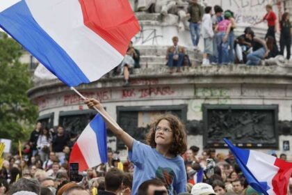 Young girl with curly hair raises a large French flag amid a crowded protest at a monument.