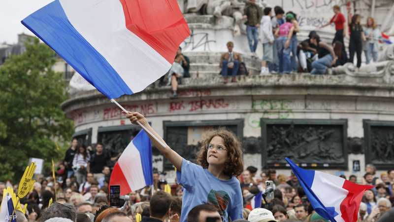 Young girl with curly hair raises a large French flag amid a crowded protest at a monument. - برفيلكس - brieflex