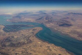 Aerial view of a blue-green river winding through a dry, desert landscape with canyons and distant mountains.