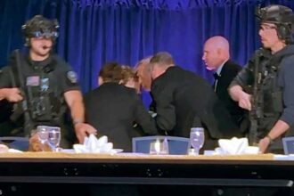 Armed security guards in tactical gear stand at a long table during a formal event, with attendees bent over in the background and blue curtains behind them.