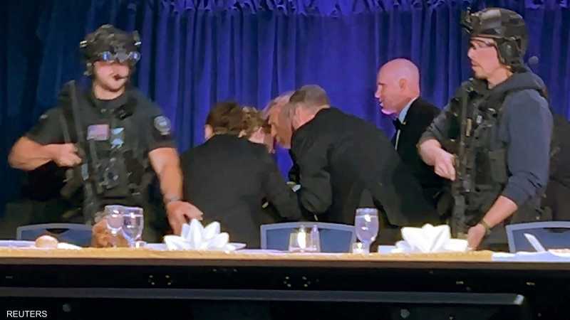 Armed security guards in tactical gear stand at a long table during a formal event, with attendees bent over in the background and blue curtains behind them. - برفيلكس - brieflex