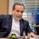 A man in a dark suit and white shirt sits at a conference table, looking thoughtful during a meeting, with a nameplate nearby. - برفيلكس - brieflex