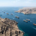 Aerial view of a narrow sea channel with numerous cargo ships sailing in a convoy along rocky desert coastline under a clear blue sky. - برفيلكس - brieflex