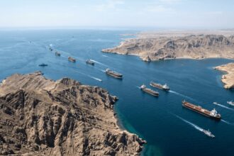 Aerial view of a narrow sea channel with numerous cargo ships sailing in a convoy along rocky desert coastline under a clear blue sky.