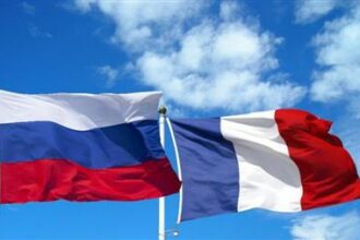 Two national flags waving in the wind: Russia on the left (white-blue-red horizontal stripes) and France on the right (blue-white-red vertical stripes) against a blue sky.