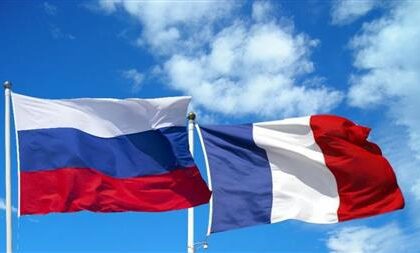 Two national flags waving in the wind: Russia on the left (white-blue-red horizontal stripes) and France on the right (blue-white-red vertical stripes) against a blue sky.