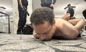 Shirtless man lying face down on a carpeted floor with several police officers standing nearby in a hallway or public space.