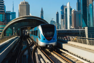 Blue and white metro train on an elevated track approaching a curved glass station, with a skyline of tall glass towers behind.