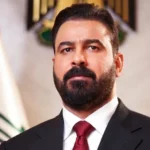 Portrait of a bearded man in a dark suit and red tie, standing in front of a flag in a formal setting. - برفيلكس - brieflex