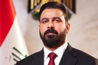 Portrait of a bearded man in a dark suit and red tie, standing in front of a flag in a formal setting.