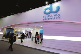 Trade show booth featuring a large blue 'du' logo and the slogan 'add life to life' on a curved purple wall, with attendees walking nearby.