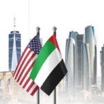 American and UAE flags flutter in front of a modern city skyline with tall skyscrapers.