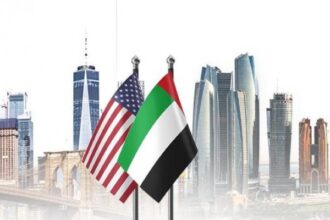 American and UAE flags flutter in front of a modern city skyline with tall skyscrapers.