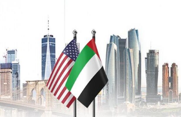 American and UAE flags flutter in front of a modern city skyline with tall skyscrapers.