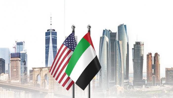 American and UAE flags flutter in front of a modern city skyline with tall skyscrapers.